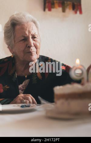 Gealterte Frau bläst Kerzen auf Geburtstagskuchen und klatscht dann die Hände, während sie den 90.. Jahrestag mit einem Verwandten zu Hause feiert Stockfoto