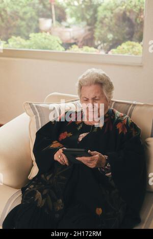 Fokussierte ältere Frau mit grauen Haaren, die auf der Couch liegt und E-Book auf dem Tablet im Wohnzimmer zu Hause liest Stockfoto