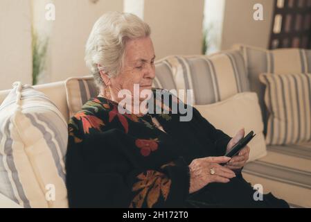 Fokussierte ältere Frau mit grauen Haaren, die auf der Couch liegt und E-Book auf dem Tablet im Wohnzimmer zu Hause liest Stockfoto