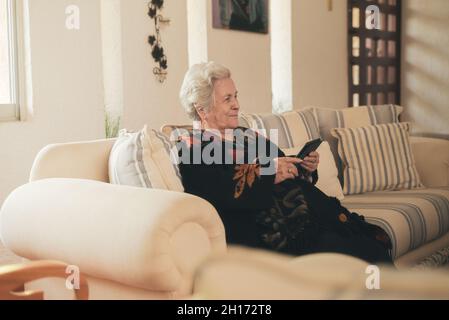 Fokussierte ältere Frau mit grauen Haaren, die auf der Couch liegt und E-Book auf dem Tablet im Wohnzimmer zu Hause liest Stockfoto