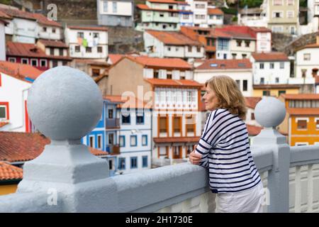 Seitenansicht der erwachsenen Reisenden, die sich am Zaun lehnen, während sie den Blick auf die typischen bunten Wohnhäuser in Asturias Stadt bewundern Stockfoto