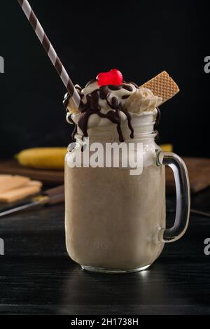 Glas mit süßem Bananenspalt-Milchshake und Schlagsahne-Waffeln Schokolade und Kirsche auf dem Tisch Stockfoto