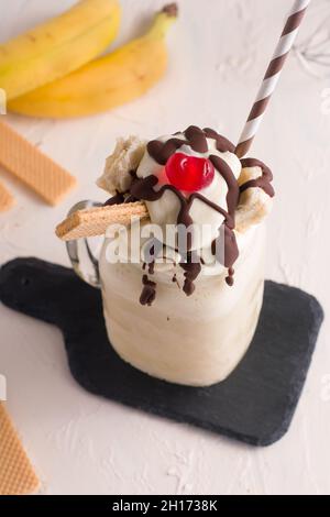 Von oben aus Glas mit süßem Bananenspalt-Milchshake, gekrönt mit Schlagsahne, Waffeln, Schokolade und Kirsche auf Schneidebrett Stockfoto