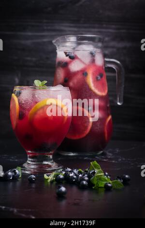 Glas und Krug mit erfrischender, kalter Limonade mit frischen Heidelbeeren und Zitronenscheiben auf einem dunklen Tisch Stockfoto