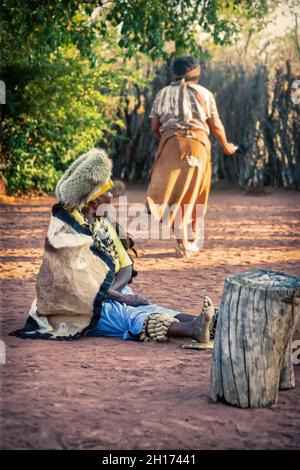 Zwei ältere Frauen aus dem südlichen Afrika, die in einer traditionellen Tracht mit Hut und Kuh gekleidet sind, verstecken sich in einem Dorf auf dem Land Stockfoto