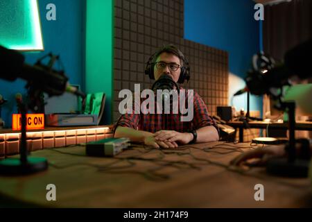 Junger bärtiger Mann Millennial in Brillen und Kopfhörern, der im Mikrofon spricht, während er Podcast im dunklen Studio aufnimmt Stockfoto