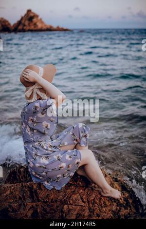 Charmante junge Frau in Sommerkleid und Hut sitzen auf felsigen Küste, während Blick weg in Sommerabend Stockfoto