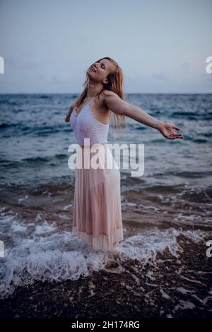 Attraktive junge Hündin in stilvoller Sommerkleidung, die mit erhobenen Armen und geschlossenen Augen am Abend im Wasser des Meeres steht Stockfoto