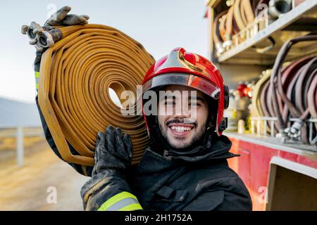 Tapferer männlicher Feuerwehrmann, der Schutzhardhut und Uniform trägt und die Kamera anschaut, während er einen großen schweren Schlauch auf der Schulter in der Nähe eines Feuerwehrwagens in Ackerland trägt Stockfoto