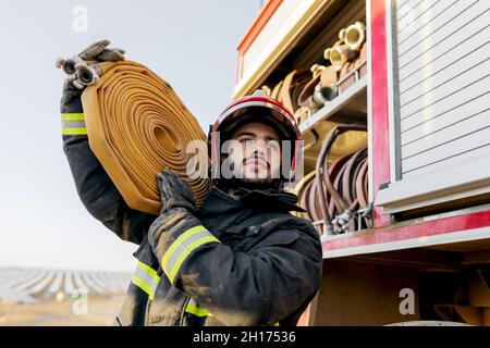 Von unten der tapfere männliche Feuerwehrmann trägt Schutzhardhat und Uniform wegschauen, während das Tragen großer schwerer Schlauch auf der Schulter in der Nähe von Feuerwehrauto Stockfoto