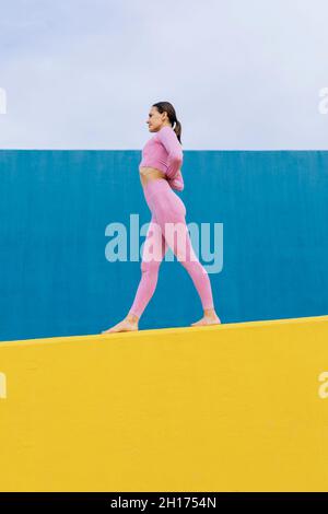 Seitenansicht der ganzen Länge einer schlanken barfuß-Frau, die Yoga im umgekehrten Gebet praktiziert, mit den Händen hinter dem Rücken an der gelben und blauen Wand Stockfoto