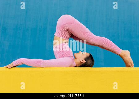 Seitenansicht einer flexiblen, schlanken Frau in Sportkleidung, die auf einer gelben Matte gegen eine hellblaue Wand trainiert Stockfoto