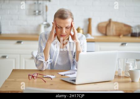 Frustrierte, reife Frau, die Kopfschmerzen hat, während sie von zu Hause aus online arbeitet Stockfoto