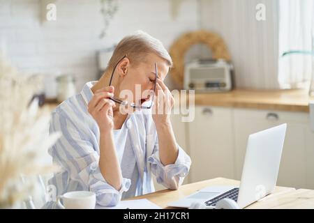 Frau mittleren Alters massiert ihre Nase und fühlt sich während der Arbeit auf Laptop online von zu Hause aus Stockfoto