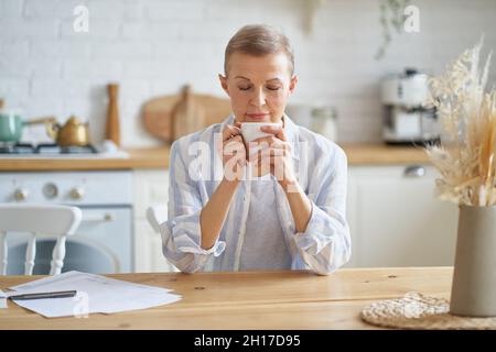 Attraktive reife Frau beginnt den Tag mit Kaffee, während sie am Holztisch in der Küche sitzt Stockfoto