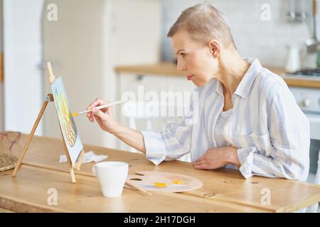 Eine Frau mittleren Alters, die zu Hause am Holztisch in der Küche Bilder malt Stockfoto