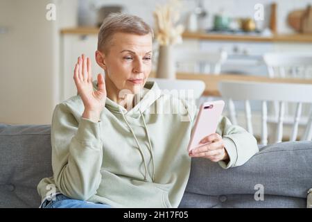 Fröhliche, fröhliche ältere Frau, die mit einem Freund oder einer Familie auf dem Smartphone einen Videoanruf führt, während sie zu Hause auf dem Sofa sitzt Stockfoto