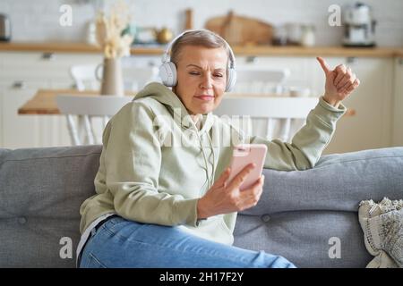 Glückliche ältere Frau mit Kopfhörern, die Musik auf dem Smartphone hört, während sie sich zu Hause auf dem Sofa entspannt Stockfoto
