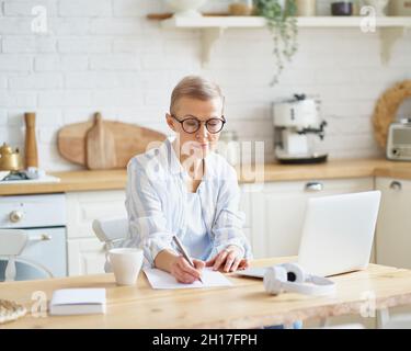 Konzentrierte ältere Schriftstellerin, die etwas auf dem Laptop tippt, während sie von zu Hause aus online arbeitet Stockfoto