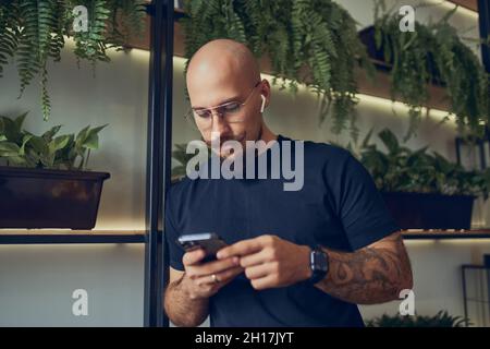 Konzentrierter Geschäftsmann nutzt das Smartphone mit Kopfhörern, während er im Heimbüro steht. Distanzjob. Stockfoto