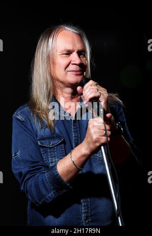 Älterer Mann mit langen grauen Haaren in blauem Denim-Hemd singt auf schwarzem Hintergrund in ein Mikrofon auf der Bühne Stockfoto