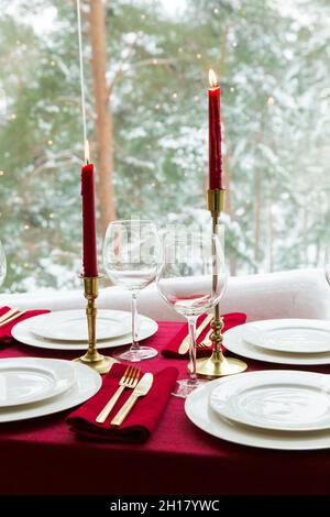 Schöne Tischkulisse mit klassischem weißem Porzellan, goldenem Besteck und Kerzen. Großes Fenster mit Winterlandschaft, Kiefernwald mit Schnee bedeckt Stockfoto