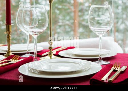 Schöne Tischkulisse mit klassischem weißem Porzellan, goldenem Besteck und Kerzen. Großes Fenster mit Winterlandschaft, Kiefernwald mit Schnee bedeckt Stockfoto