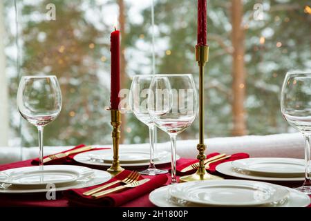 Schöne Tischkulisse mit klassischem weißem Porzellan, goldenem Besteck und Kerzen. Großes Fenster mit Winterlandschaft, Kiefernwald mit Schnee bedeckt Stockfoto