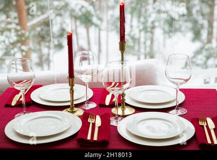 Schöne Tischkulisse mit klassischem weißem Porzellan, goldenem Besteck und Kerzen. Großes Fenster mit Winterlandschaft, Kiefernwald mit Schnee bedeckt Stockfoto