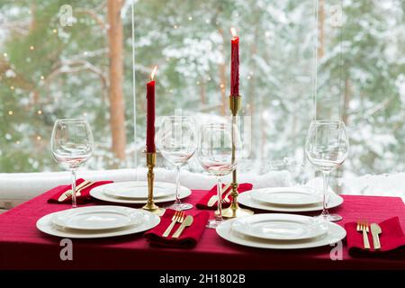 Schöne Tischkulisse mit klassischem weißem Porzellan, goldenem Besteck und Kerzen. Großes Fenster mit Winterlandschaft, Kiefernwald mit Schnee bedeckt Stockfoto