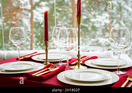 Schöne Tischkulisse mit klassischem weißem Porzellan, goldenem Besteck und Kerzen. Großes Fenster mit Winterlandschaft, Kiefernwald mit Schnee bedeckt Stockfoto