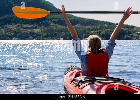 Das Mädchen hält ein Paddel über ihren Kopf und schwimmt entlang des Fjords in norwegen, Kajakfahren im Meer, Outdoor-Aktivitäten Stockfoto