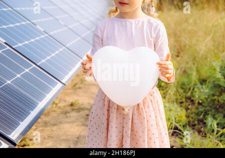 Unkenntlich Mädchen Kind mit weißen leeren Herz geformten Ballon, neben Sonnenkollektoren stehen, Solarfarm an sonnigen Sommertag. Förderung grüner Energie Stockfoto