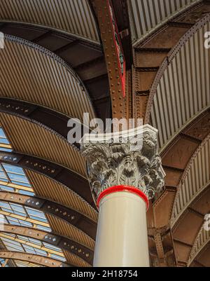 Blick nach oben auf eine historische korinthische Säule aus dem 19. Jahrhundert, die einen Baldachin stützt. Ein Oberlicht wirft Licht auf ein kompliziertes Eisenwerk. Stockfoto