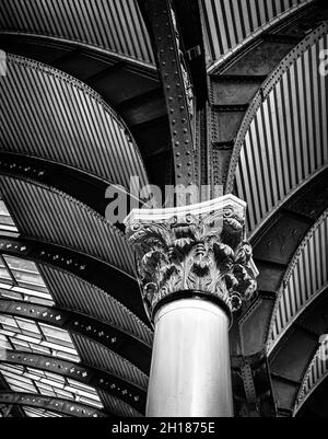 Blick nach oben auf eine historische korinthische Säule aus dem 19. Jahrhundert, die einen Baldachin stützt. Ein Oberlicht wirft Licht auf ein kompliziertes Eisenwerk. Stockfoto