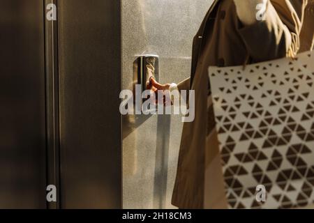 Nahaufnahme eines Fingers drückt die Aufzugstaste. Eine Frau mit Einkaufstaschen in beigem Regenmantel ruft den Aufzug an. Ober- oder Untergeschoß Stockfoto