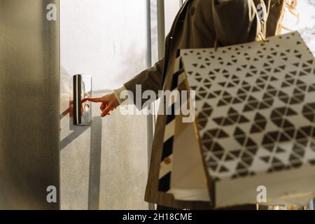 Nahaufnahme eines Fingers drückt die Aufzugstaste. Eine Frau mit Einkaufstaschen in beigem Regenmantel ruft den Aufzug an. Ober- oder Untergeschoß Stockfoto