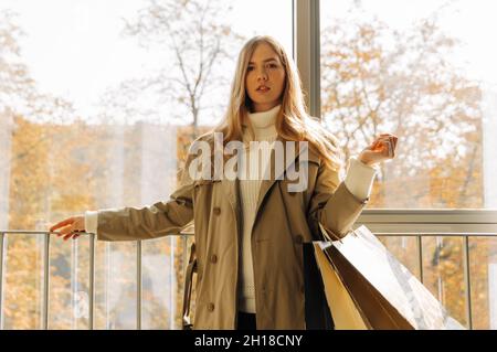 Frau im beigen Regenmantel geht im Herbst mit Einkaufstaschen in den Händen durch das Einkaufszentrum. Konzept: Herbstverkauf, Rabatte. Auf gegen sh Stockfoto