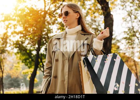 Stilvolle Frau in einem beigen Regenmantel und Sonnenbrillen mit Einkaufstaschen in den Händen, auf gegen die Herbstbäume, Saison der Rabatte, Verkauf. Business wo Stockfoto