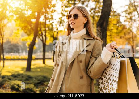 Stilvolle Frau in einem beigen Regenmantel und Sonnenbrillen mit Einkaufstaschen in den Händen, auf gegen die Herbstbäume, Saison der Rabatte, Verkauf. Business wo Stockfoto