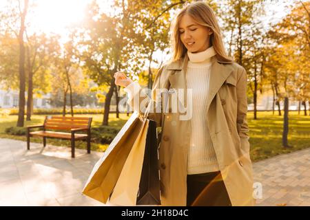 Nahaufnahme einer stylischen Frau in beigem Regenmantel und Sonnenbrillen mit Einkaufstaschen in den Händen, auf gegen die Herbstbäume, Saison der Rabatte, Sal Stockfoto