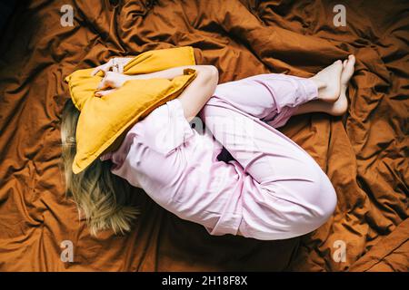 Depressive kaukasische tausendjährige Frau, die allein auf dem Bett lag und den Kopf mit Kissen bedeckte, die Angst hatten, Draufsicht. Stockfoto