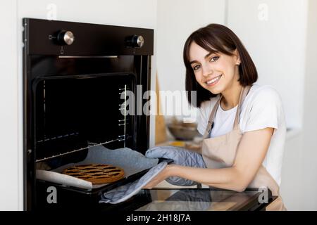 Porträt einer glücklichen Dame, die in der Küche backt, Schürze und Fäustling trägt und das Blech mit hausgemachtem Kuchen aus dem heißen Ofen nimmt Stockfoto