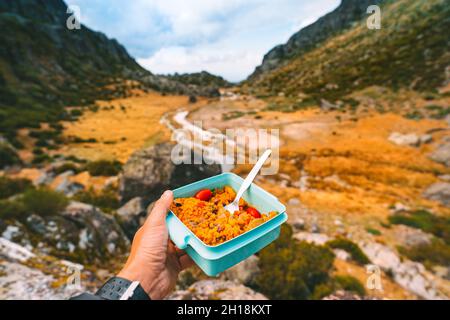 Hand des Mannes hält Kunststoff-Lebensmittelbehälter mit gesunden hausgemachten Lebensmitteln. Mittagessen beim Wandern in den Bergen Stockfoto