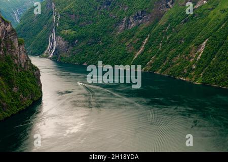 Ein Bootsruck kräuselt durch den Geirangerfjord, der sich durch waldreichen, steilen Klippen und Bergen schlängelt, Seven Sisters Wasserfälle liegen in der Ferne. Geira Stockfoto