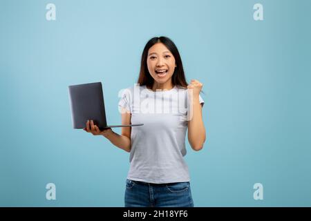 Überglücklich asiatische Dame Anheben geballte Faust und feiert Erfolg mit Laptop-Computer, stand auf blauem Hintergrund Stockfoto