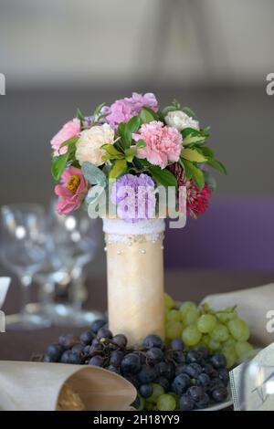 Blumenarrangement im Mittelpunkt mit pastellfarbenen Nelken in Elfenbeinvase mit Perlenperlen und frischen weißen und roten Trauben in der Nähe Stockfoto