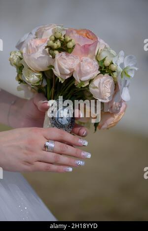 Nahaufnahme der kaukasischen Braut mit makelloser Maniküre mit einem runden Pastellstrauß mit einer Vintage-Cameo-Brosche mit großen weißen Orchideen Stockfoto