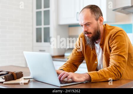 Mann verwenden Laptop für Online-Ausbildung oder Einkaufen. Ruhiger junger Mann, der von zu Hause aus arbeitet. Porträt eines kühnen, bärtigen kaukasischen Mannes, der seinen Freiberufler verwendet Stockfoto