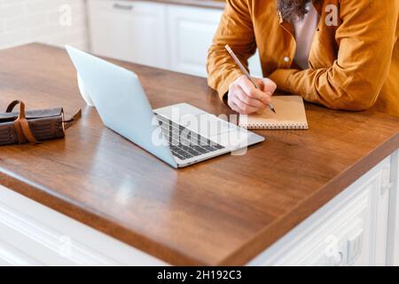 Mann machen Papierkram mit Laptop für Online-Arbeit Ausbildung oder Arbeit von zu Hause aus. Nahaufnahme Mann mit männlichen Händen schreibt mit Bleistift schreibt Notizen in Notizblock Stockfoto